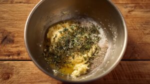A mixing bowl with butter, chopped herbs, garlic powder, salt, and pepper on a wooden surface, ready to be combined