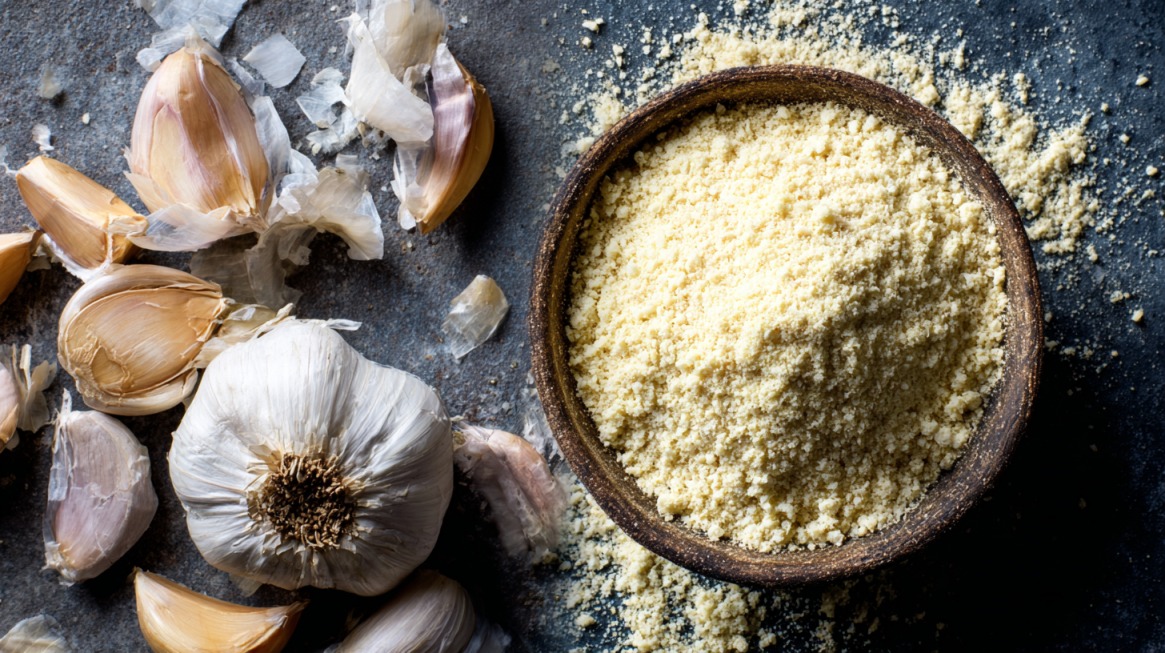 Garlic powder in a wooden bowl next to scattered garlic cloves and peels on a dark surface
