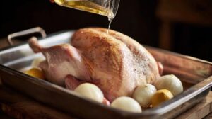 Raw whole turkey in a roasting pan with onions and lemon, being drizzled with oil or melted butter