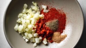 Chopped sautéed onions and a variety of chili spices in a bowl, ready to be mixed