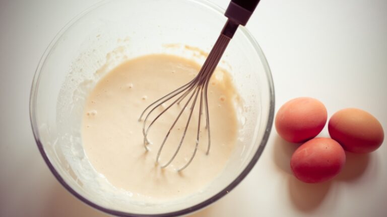 A bowl of raw pancake batter with a whisk and fresh eggs nearby