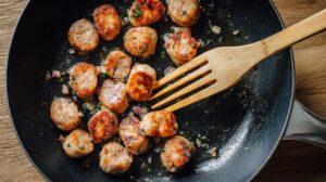 Sliced sausage pieces being cooked in a skillet with onions and herbs, stirred with a wooden spatula
