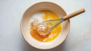 A mixing bowl with whisked eggs, milk, and seasonings being combined with a wire whisk