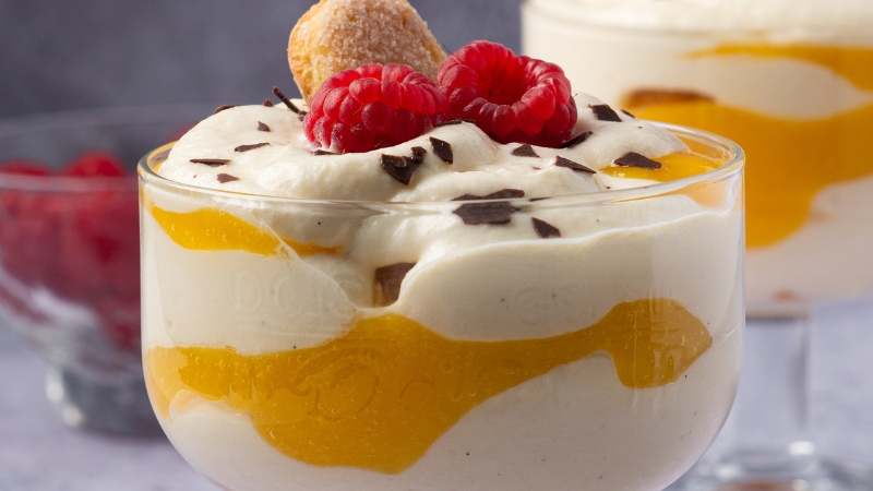 A dessert glass filled with creamy layers, mango puree, raspberries, chocolate shavings, and a biscuit garnish