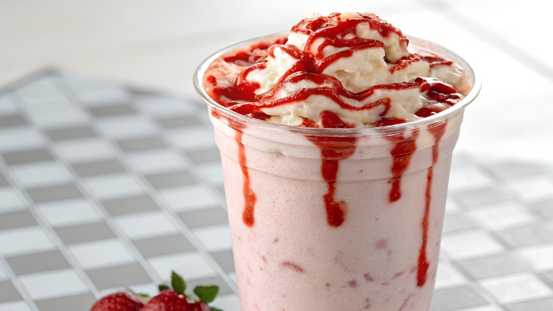 A clear dessert glass filled with strawberry milkshake, topped with whipped cream and strawberry sauce