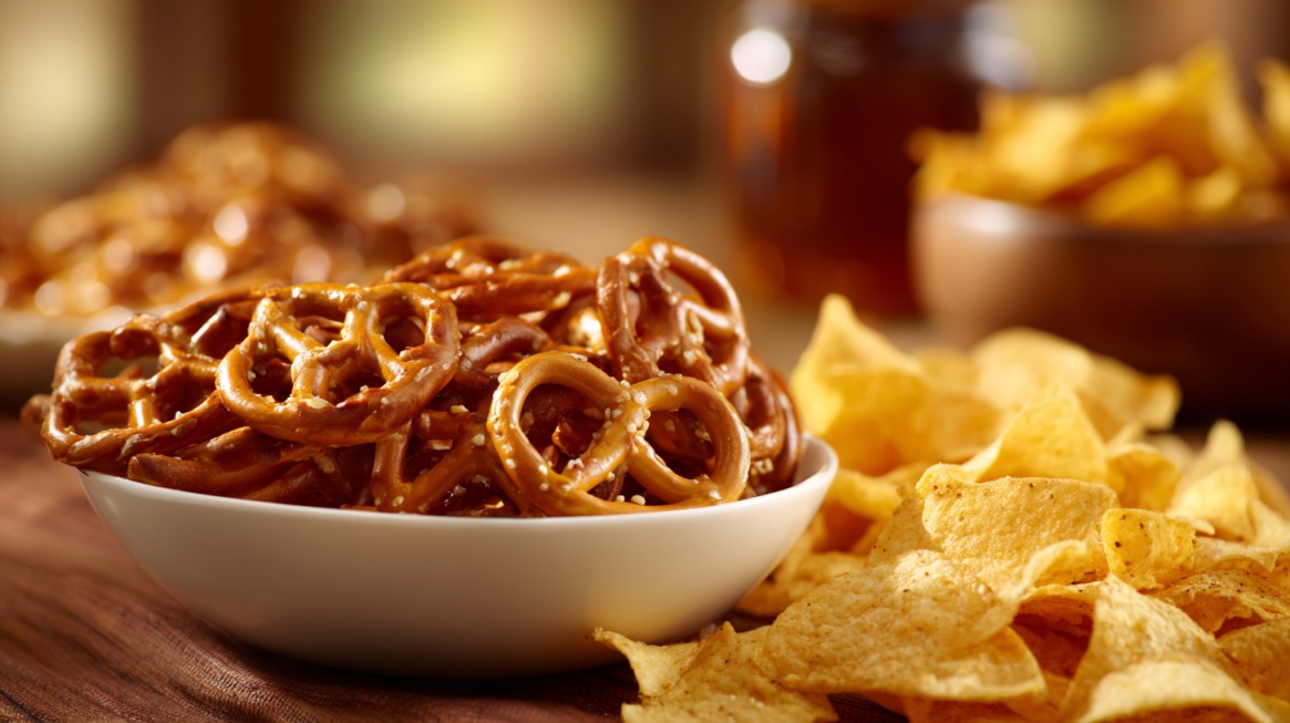 Close-up of a bowl filled with small salted pretzels surrounded by potato chips on a wooden table in warm lighting