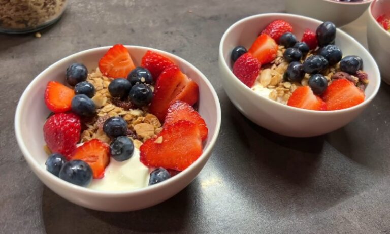 Two white bowls filled with yogurt, granola, sliced strawberries, and whole blueberries on a dark countertop