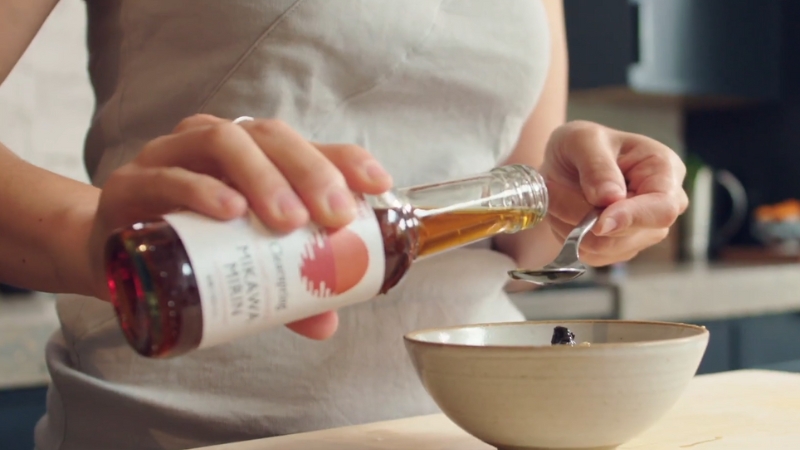 A person pours mirin from a bottle into a spoon over a bowl in a kitchen