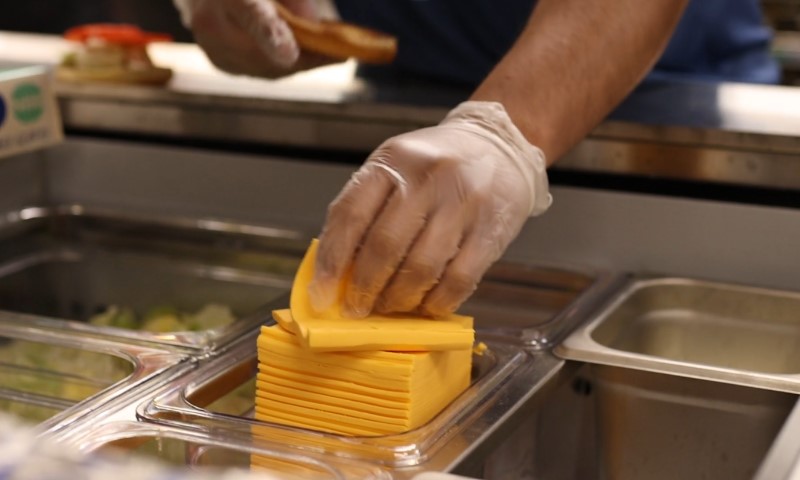 Close-up of a gloved hand stacking vibrant orange cheddar cheese slices in a stainless steel food service tray