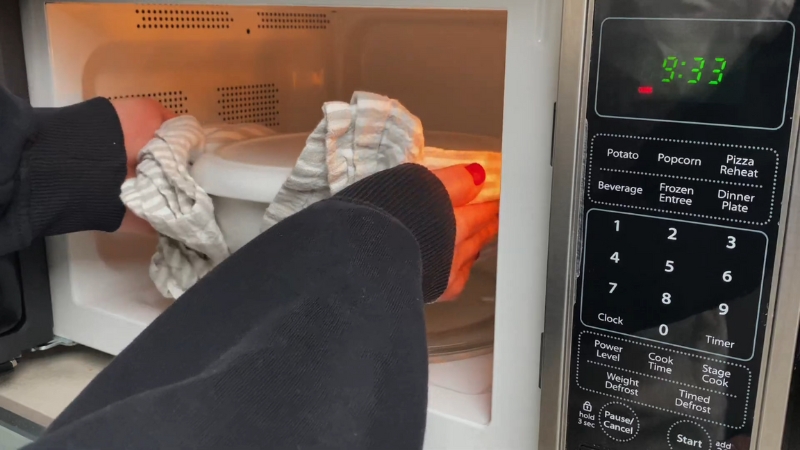Hands placing a covered food container into a microwave to reheat leftovers evenly