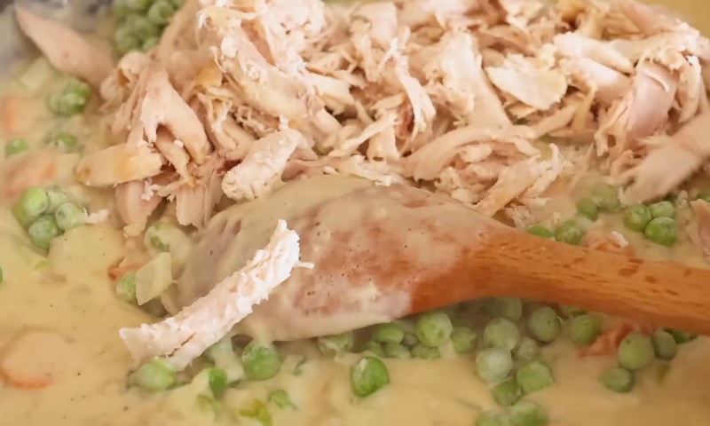Shredded chicken, peas, and carrots are being mixed into a thick creamy sauce with a wooden spoon