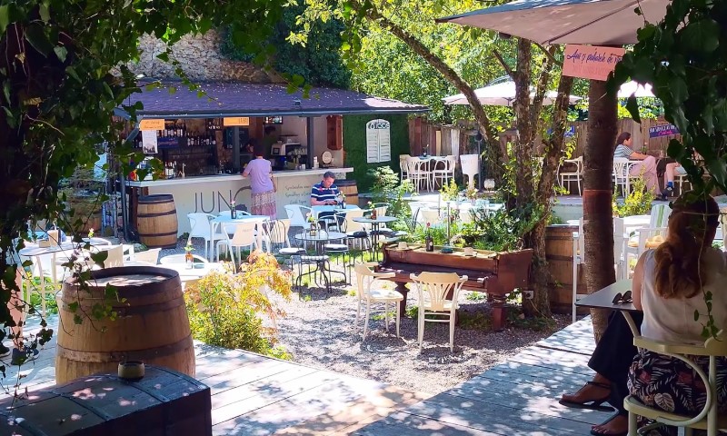 Outdoor café with lush greenery and dappled sunlight. Patrons sit at white tables, enjoying drinks