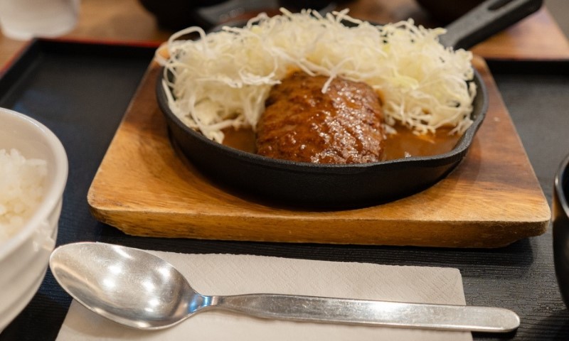 A sizzling beef patty in a cast iron skillet, topped with shredded cabbage, sits on a wooden board