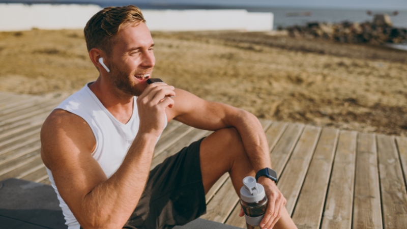 Man eating a protein bar outdoors after a workout while holding a shaker bottle