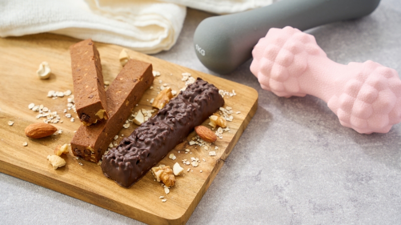 Protein bars with nuts and oats on a board next to small dumbbells for fitness use