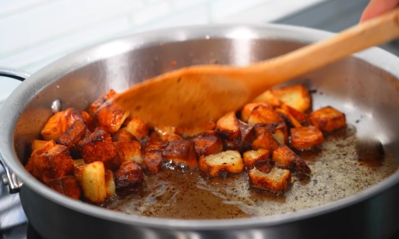 A skillet with sizzling, golden-brown roasted potatoes is being stirred with a wooden spatula