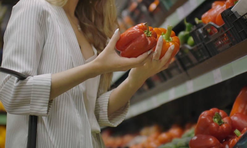 Young woman in a striped blazer selects vibrant red bell peppers in a well-lit grocery store, conveying a sense of freshness and careful choice